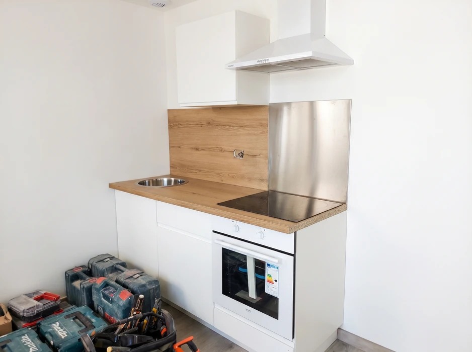 Petite cuisine compacte en cours d'installation, équipée d'un plan de travail en chêne, d'un évier rond encastré inox, d'une plaque de cuisson vitrocéramique, d'un four encastrable blanc et d'une hotte aspirante murale blanche