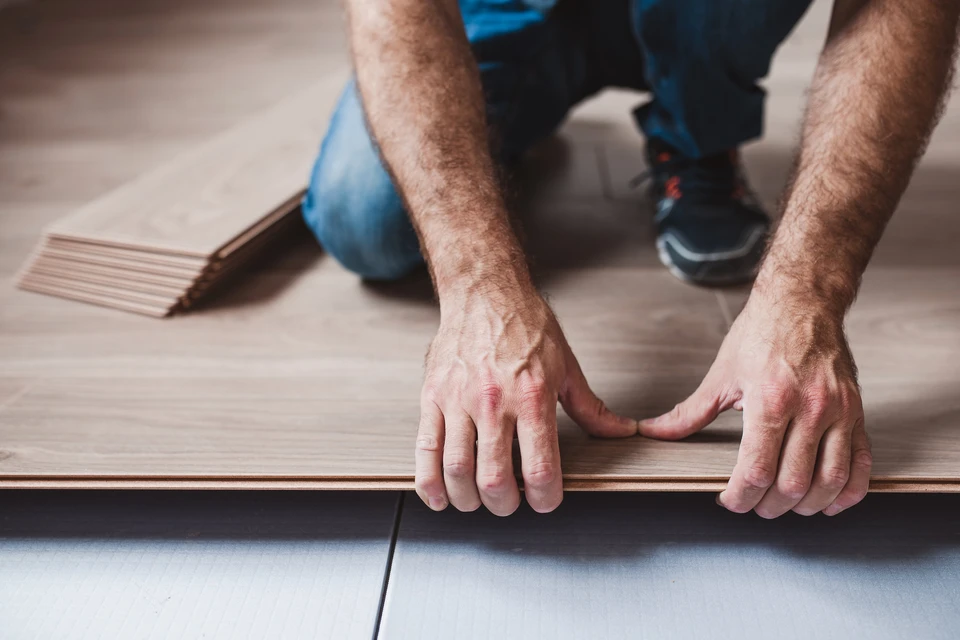 Deux artisans travaillant côte à côte sur un chantier de rénovation intérieure, l'un posant du parquet en bois tandis que l'autre effectue une tâche complémentaire à proximité ou supervise. Scène dans un intérieur résidentiel en cours de rénovation, montrant clairement la collaboration et l'implication conjointe des deux professionnels sur le terrain.